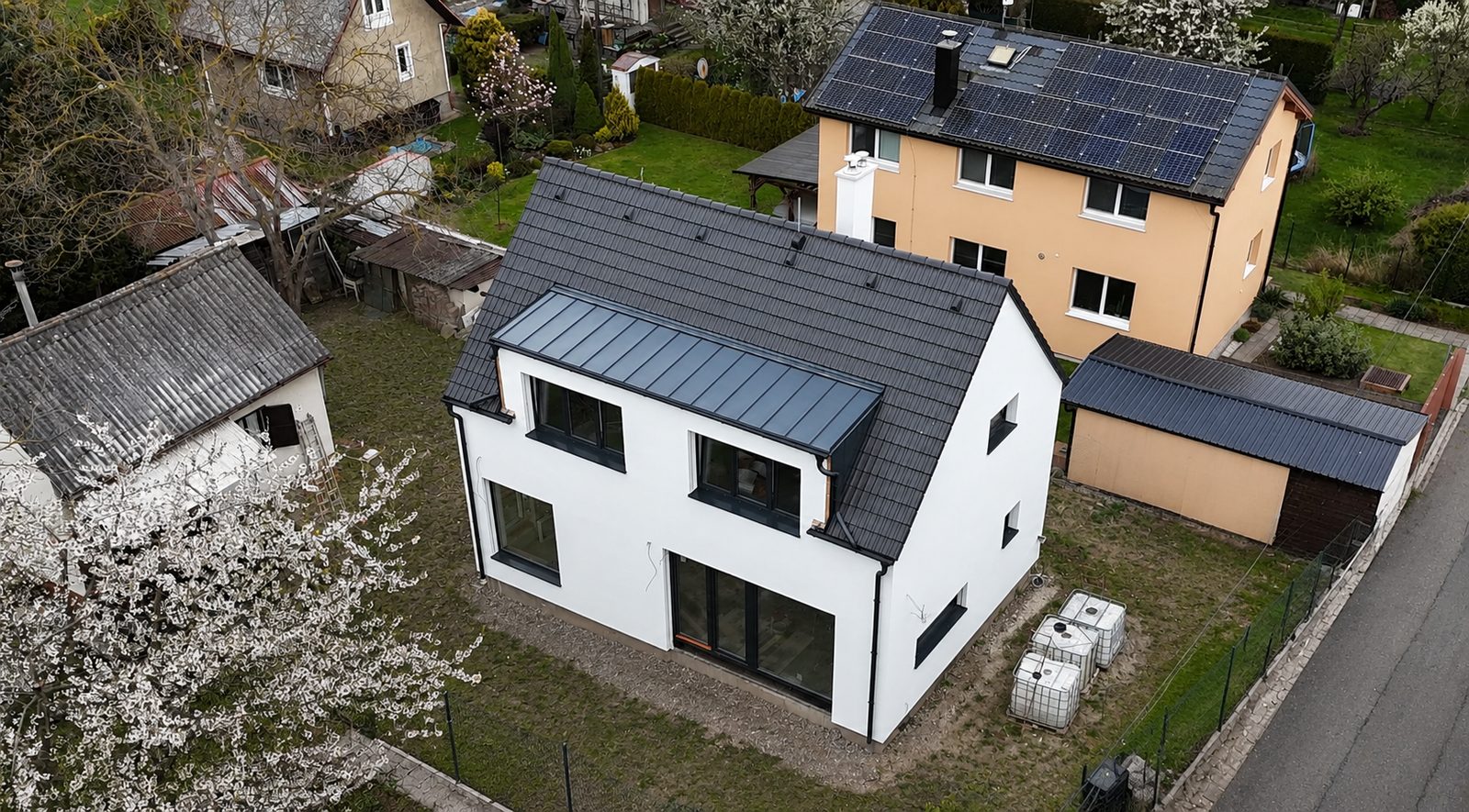 Realizace - bílý patrový dům s tmavou plechovou střechou, letecký pohled